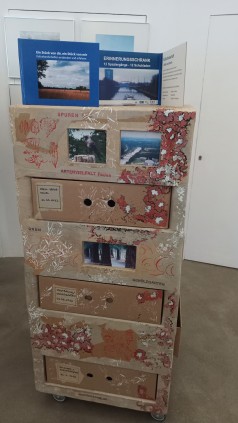 Ein Bunter Schrank mit Schubladen an allen vier Seiten. Oben drauf steht ein aufgeklappter Bildband