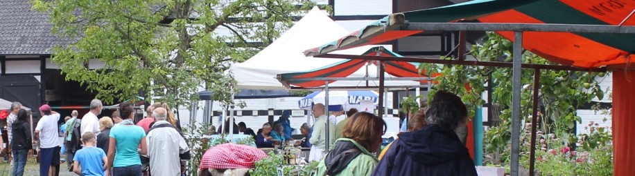 Marktstände stehen vor einem Fachwerkhof. Menschen laufen über einen Weg und stehen an den Ständen. Der Himmel ist grau.